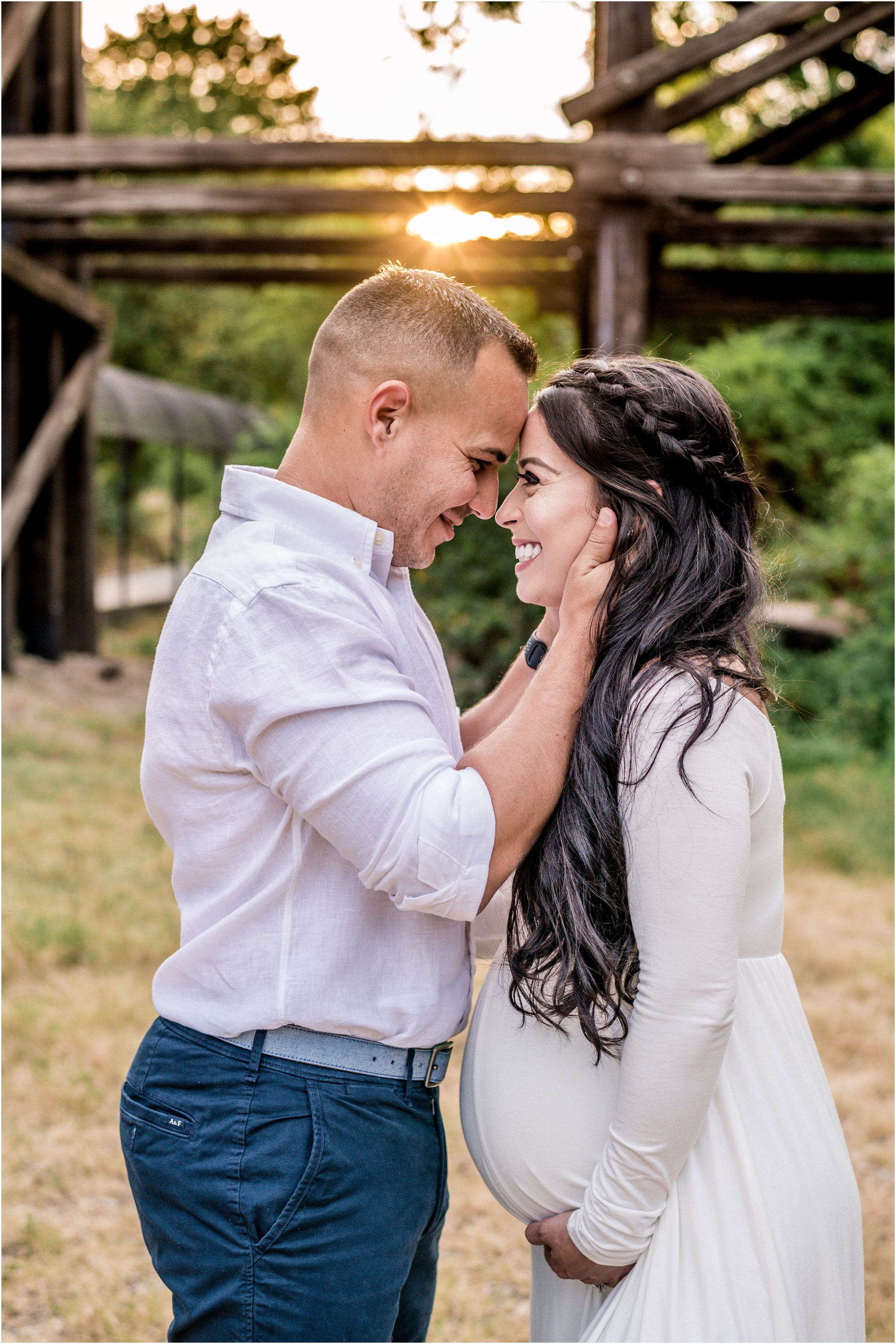 Beautiful couple giggling with one another during maternity photoshoot at sunset golden hour by bridge in Cedar Park Texas in white maxi dress natural light family photographer