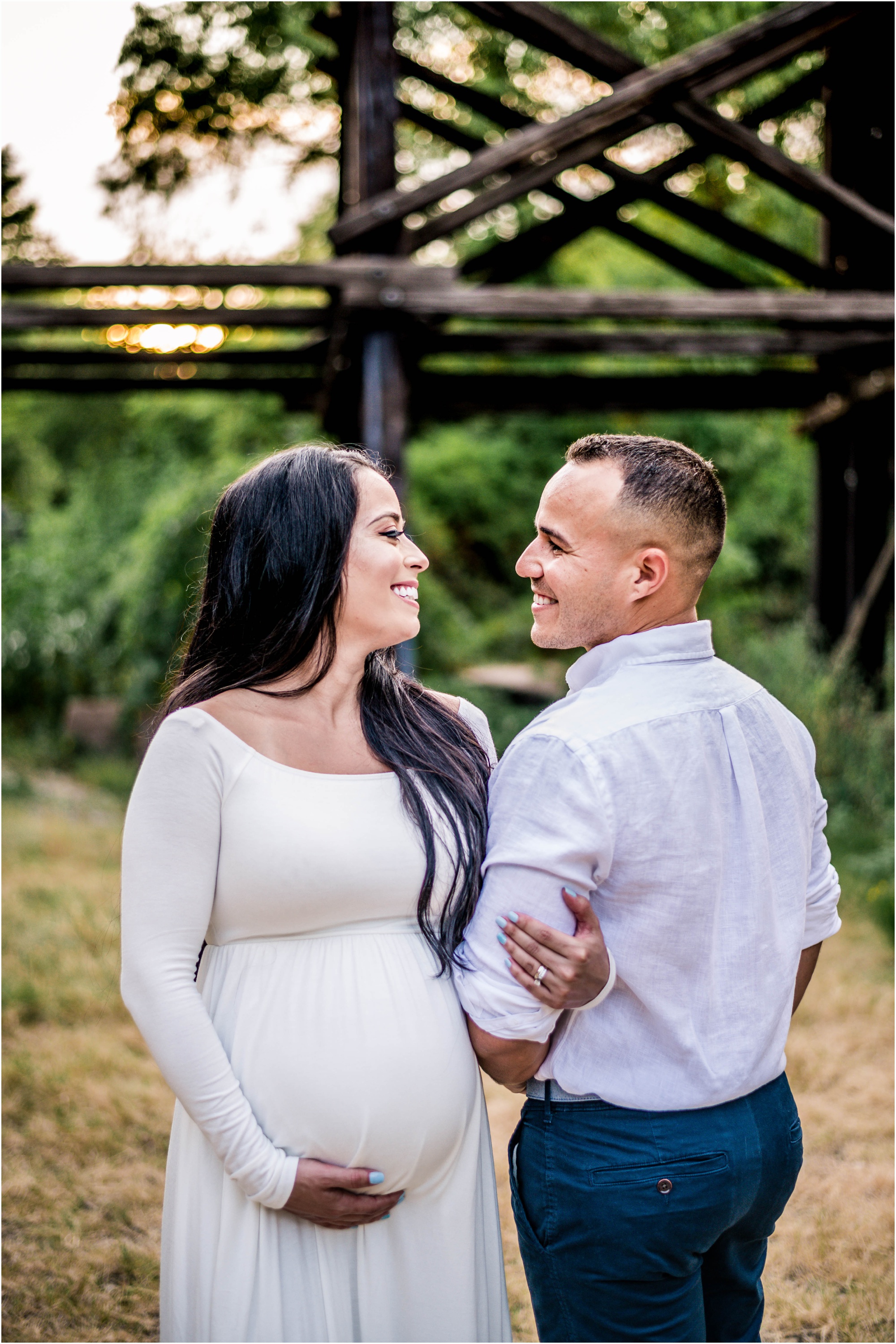 Beautiful couple giggling with one another during maternity photoshoot at sunset golden hour by bridge in Cedar Park Texas in white maxi dress natural light family photographer